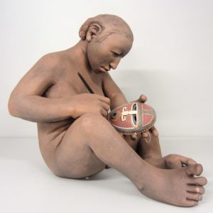 A clay sculpture of a person sitting and painting a mask.