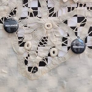 Artwork from Knowing Her CSU exhibition featuring ivory lace tablecloth against dark backing. Two black pins with the word 'RESILIENCE' are attached to the lace, which has small circular buttons throughout. Made from Deloris' tablecloth, wood, buttons, and vinyl.