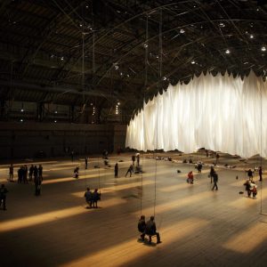 An overhead shot of an art exhibition featuring a large expanse of white cloth and people standing and sitting in front of it.
