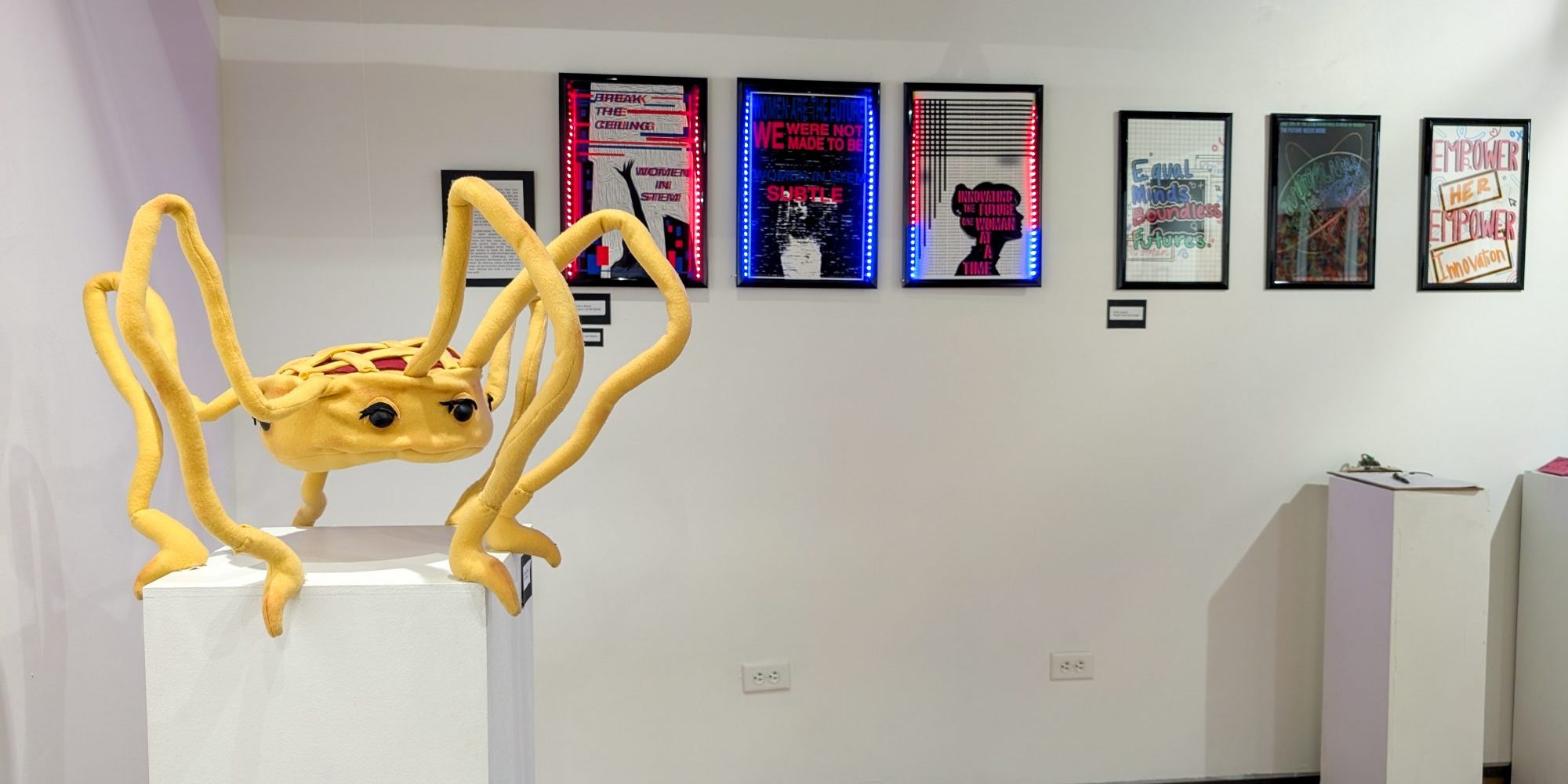 A plush sculpture of a pie with spider-like legs on a white pedestal, with framed artwork displayed on the wall behind it.