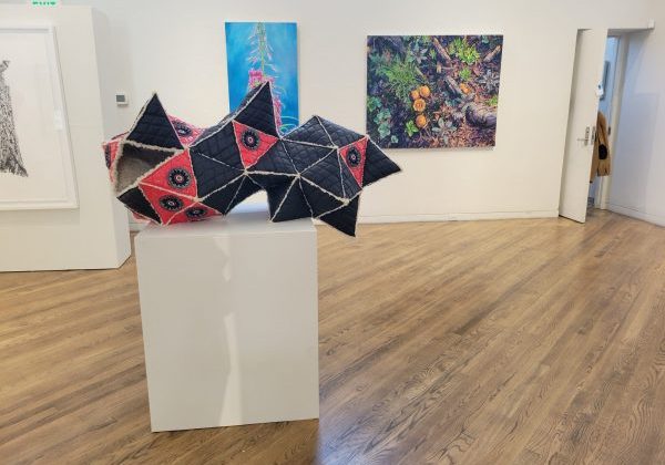 Geometric quilted sculpture on a white pedestal in a gallery with paintings on the walls and wood flooring.