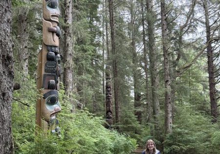young woman stands in pine forest alongside totem pole